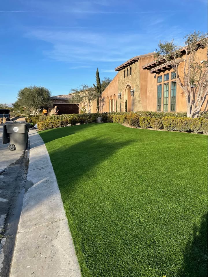 A well-maintained green lawn, created by expert artificial turf installers, borders a large tan house with tall windows under a clear blue Fountain Hills AZ sky. A sidewalk and trash bins are visible on the left side.