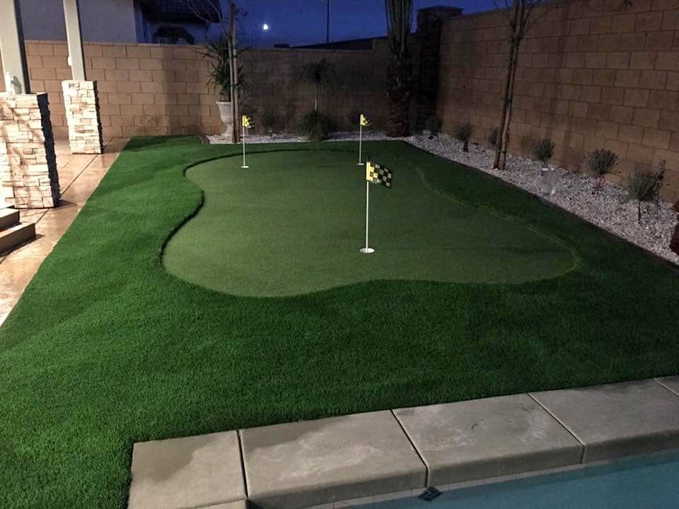 A small backyard putting green with three golf holes, each marked by a yellow flag, features synthetic golf greens surrounded by artificial grass lawns and a concrete patio near a pool and stone wall at dusk.
