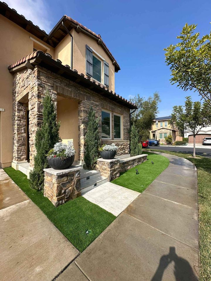 Two-story house with stone and stucco exterior in Fountain Hills AZ, featuring a small manicured lawn—perfect for artificial grass installation—potted plants by the entrance, and a sidewalk curving around the front in a sunny suburban neighborhood.