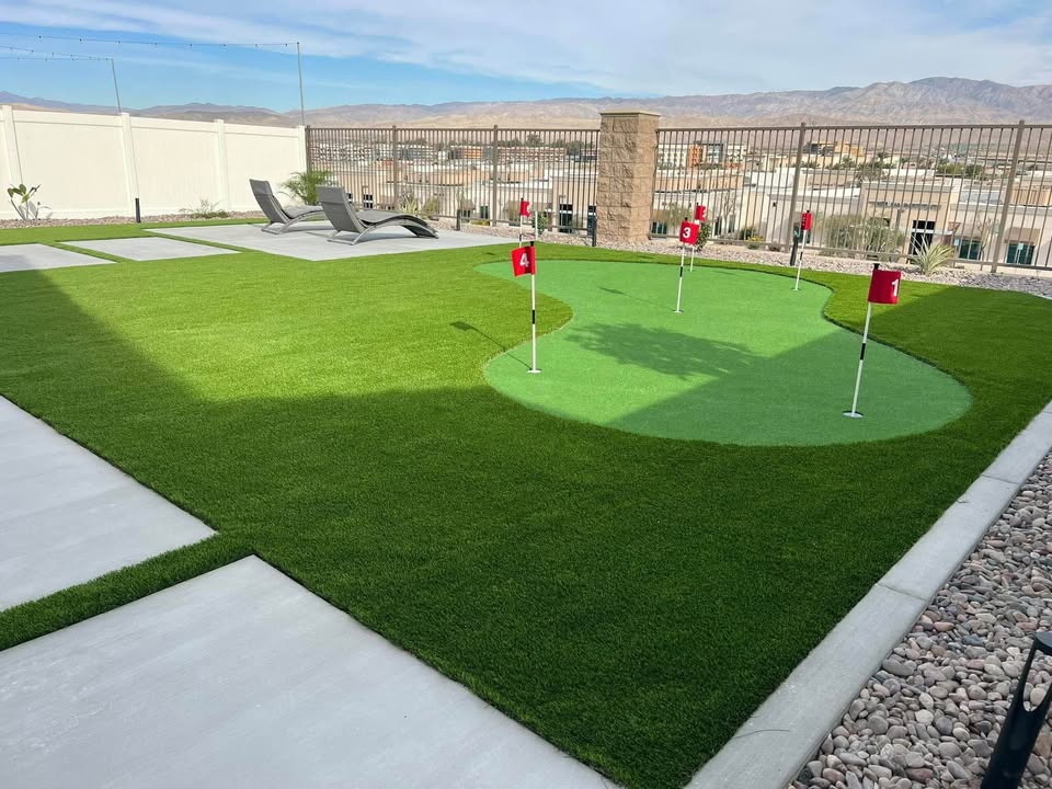 A backyard in Fountain Hills AZ showcases expert artificial grass installation with a putting green, four red flags, lounge chairs on a concrete pad, and scenic views of houses and mountains beyond a metal fence.