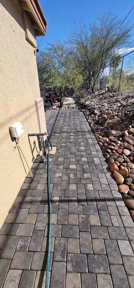 A paved walkway runs alongside a beige house with a green garden hose on the ground. On the right, decorative rocks and desert plants showcase expert Fountain Hills hardscaping under a clear blue sky.