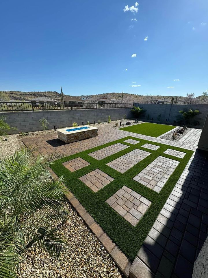 A backyard in Fountain Hills with geometric paver installation and artificial grass patterns, a raised rectangular stone firepit, desert landscaping, gravel, and a block wall fence under a clear blue sky. Desert hills are visible in the background.
