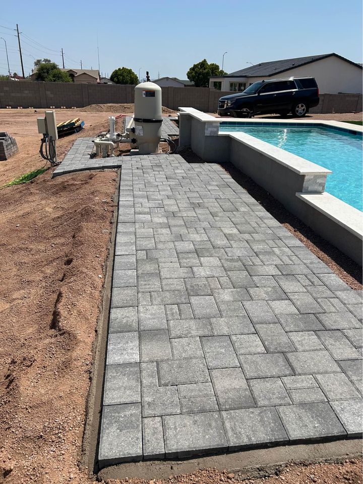 Gray stone pavers, expertly installed as part of a Fountain Hills hardscaping project, form a walkway beside a swimming pool. Nearby pool equipment and dirt outline the exterior landscape, while a black SUV sits by the house under a clear sky.