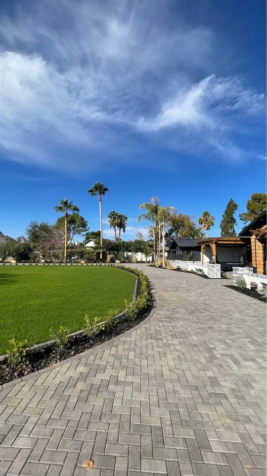 A wide stone-paved pathway, crafted with expert paver installation, curves beside a green lawn bordered by small plants, with palm trees, houses, and a blue sky with wispy clouds completing the exterior landscape.