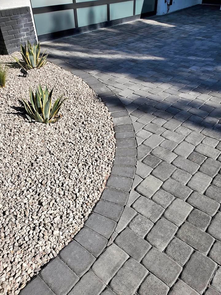 Curved grey stone pavers border a bed of small, light-colored rocks and spiky green plants outside a modern building—perfect inspiration for artificial grass installation in Fountain Hills AZ.