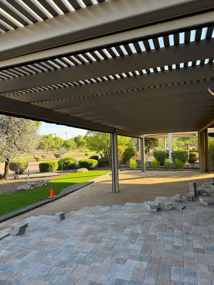 A covered patio in Fountain Hills AZ features partially completed tile flooring, stacked tiles, and a landscaped yard with bushes and trees. A single orange cone sits on the dirt beside the tiled area, highlighting ongoing hardscaping work.