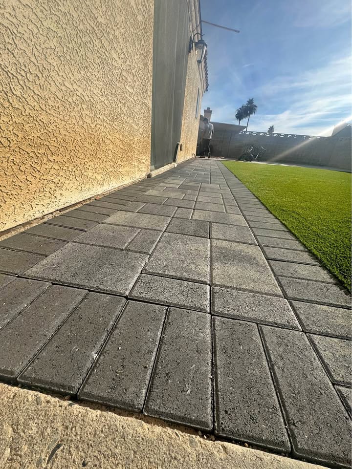 Paved stone walkway, a result of expert paver installation, runs alongside a stucco house wall, bordered by artificial green grass on the right, under a sunny Fountain Hills sky with a few trees visible in the background.