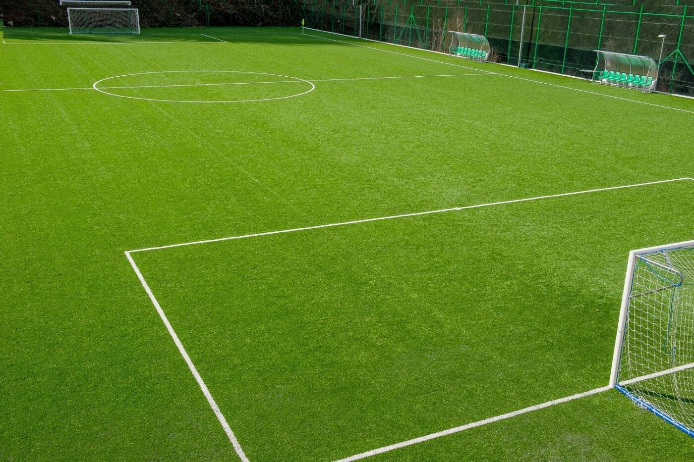 A view of an empty outdoor soccer field in Fountain Hills showcases bright green artificial grass from recent artificial turf installation, crisp white boundary lines, two goals, benches, and a surrounding green fence.