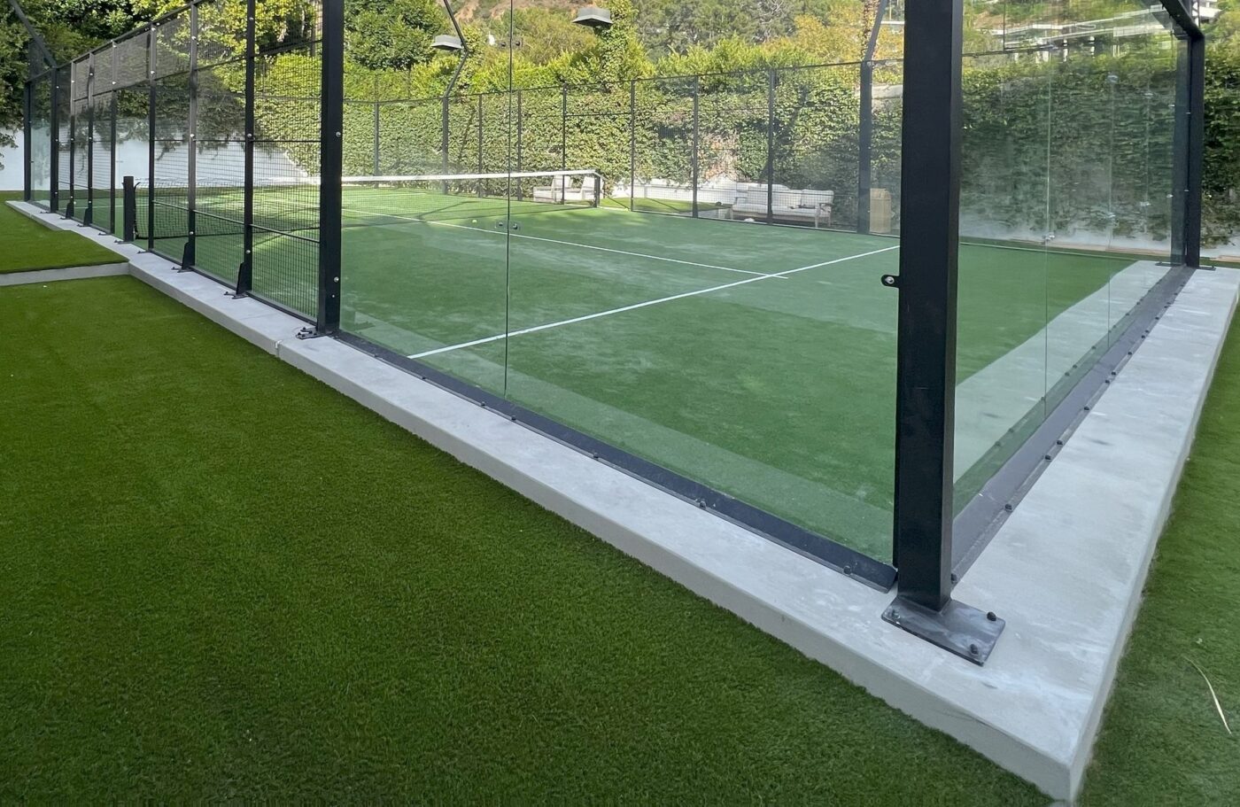 A modern outdoor padel court with glass walls and green artificial turf installation, surrounded by a hedge and benches in the background. The court in Fountain Hills is enclosed by a metal frame and set on a concrete base.