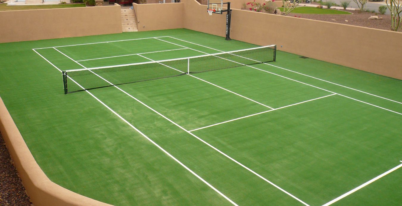 A green backyard sports court with white lines for both tennis and pickleball features artificial grass, surrounded by tan stucco walls, with a basketball hoop at one end and desert landscaping visible outside in scenic Fountain Hills.