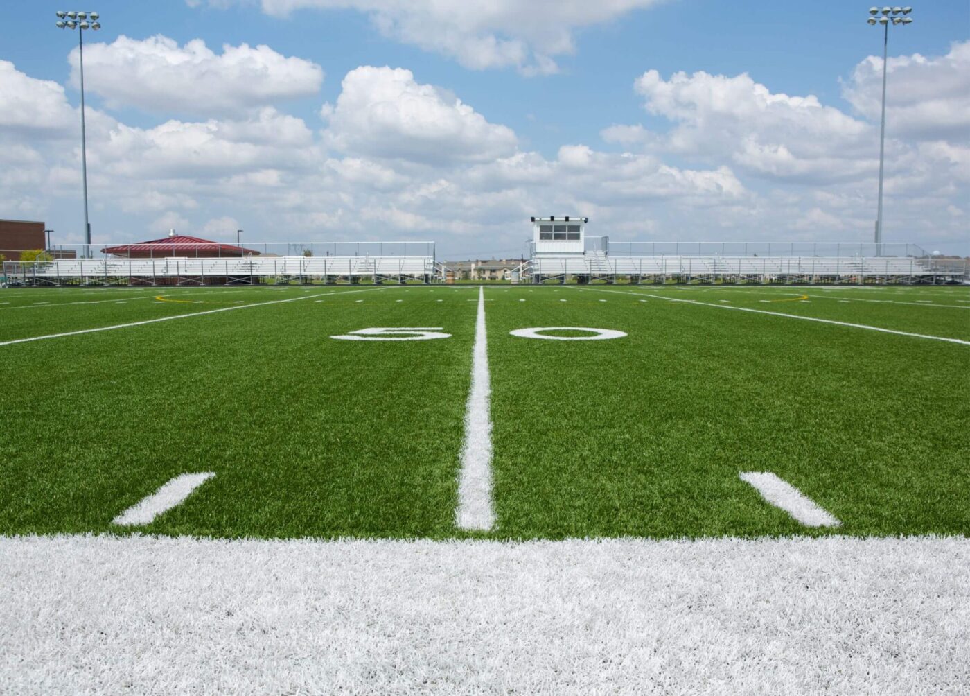 A football field viewed from the 50-yard line, with bright green turf resembling artificial grass lawns, white yard markings, bleachers, and a press box in the background under a partly cloudy sky.