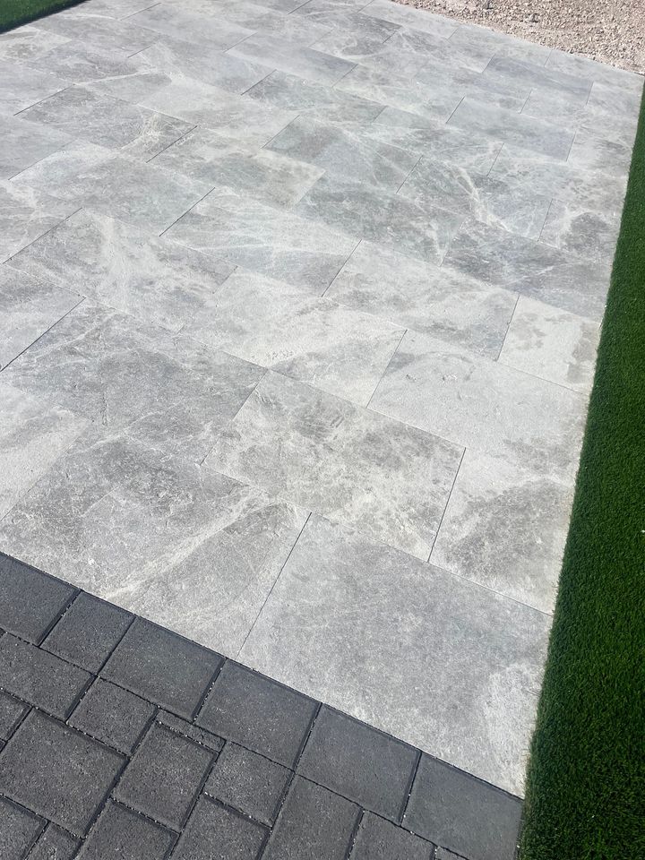 A beautifully hardscaped patio in Fountain Hills, featuring large light gray rectangular stone paver installation bordered by dark bricks, with artificial green grass and small gravel stones on either side.