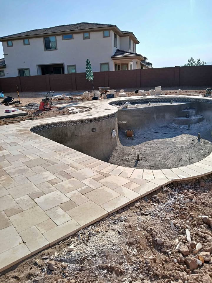 A backyard in Fountain Hills AZ under construction with an unfinished in-ground swimming pool, new paver installation, scattered tools, and a two-story house in the background, showcasing ongoing hardscaping work.