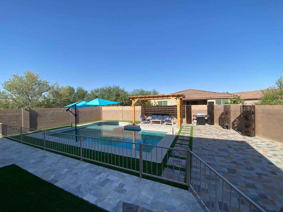 A backyard with a swimming pool, a paved patio featuring expert paver installation, lounge chairs, a blue umbrella, pergola seating, and fencing. The exterior landscape includes a single-story house and trees under a clear blue sky.