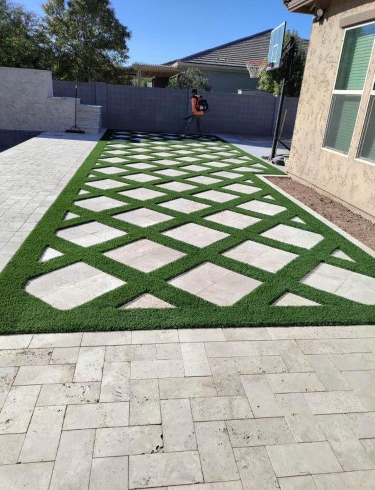 Backyard with a patio featuring a diamond-patterned design of green artificial grass and concrete pavers; a person with a leaf blower stands near the back fence, showcasing expert artificial grass installation in Fountain Hills AZ.