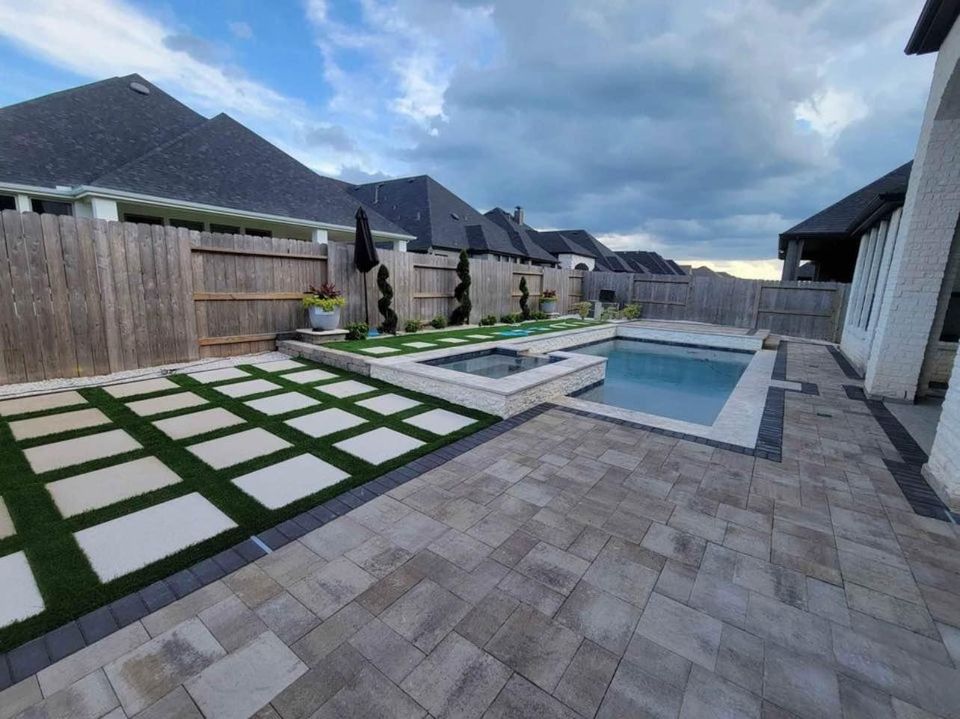 Modern backyard with a rectangular pool and attached hot tub, featuring expert paver installation and stone tiles. The patio's square pavers and artificial grass create a stylish exterior landscape, bordered by a wooden fence and neighboring houses.